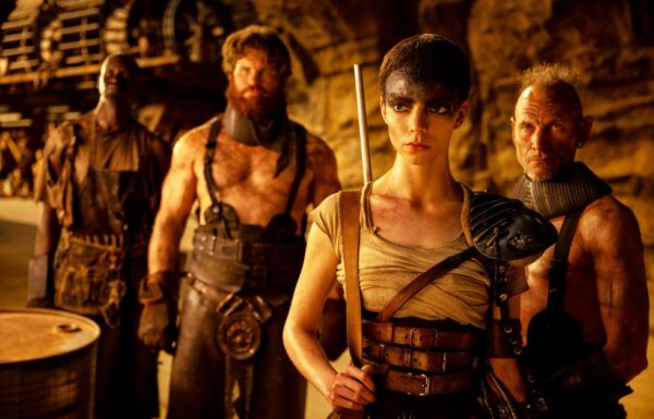 Short haired Furiosa (Anya Taylor-Joy) stands in front of a man with a bear and overalls