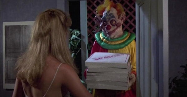 A Klown delivers a stack of pizza boxes to a scantily clad blonde