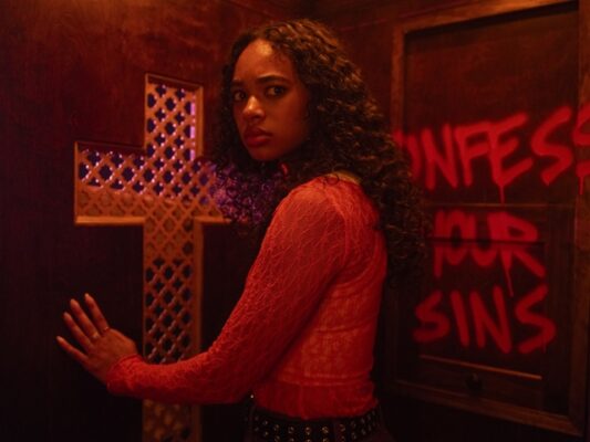 Tabby (chandler-kinney) stands in a confessional bathed in red light