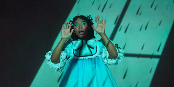 Delaney Hayles as Claudia, in a blue dress, with her hands raised on stage