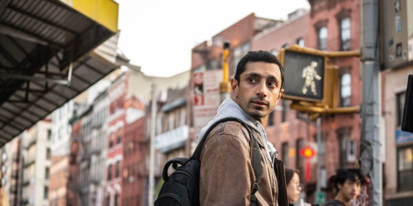 Riz Ahmed as Ash, wearing a grey hoodie, tan jacket and black backpack