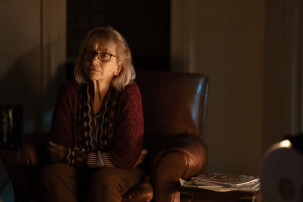 Ellen (Kathy Baker) sitting in a brown armchair in a red sweater and glasses