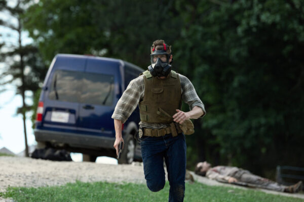 Olsen (Bill Heck) running away from a van, wearing body armor and a gas mask