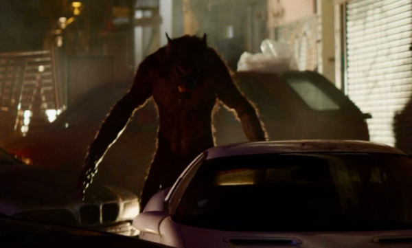 A long shot of a werewolf standing partially in shadow between cars at night