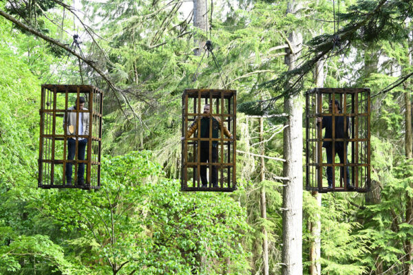 Three men hang in enormous cages suspended in the air in the woods