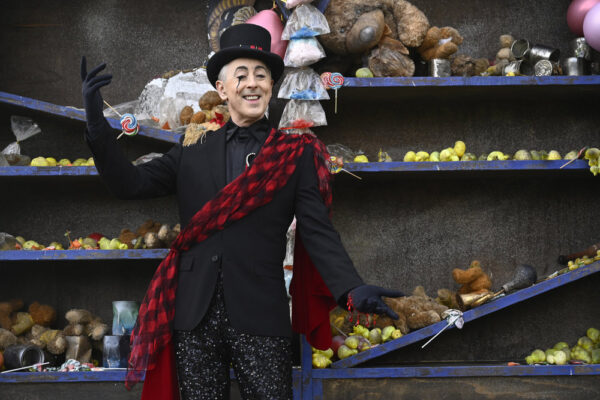 Alan Cumming in a black suit, top hat and checkered red and black scarf draped across his chest, in front of a row of carnival toys