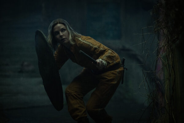 A woman in an orange jumpsuit crouches, clutching a makeshift shield