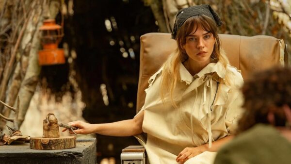 A seated teen girl wrapped in a white shawl and blue bandana reaches for a wooden gavel on a tree stump