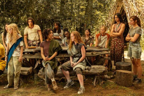 A group of teen girls in tattered garb sit and stand around a makeshift wooden table in the woods