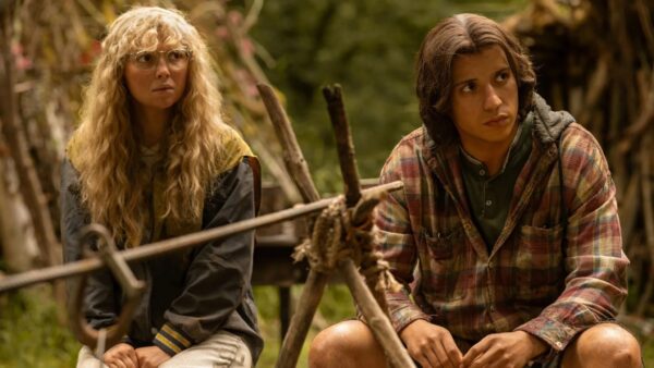 A blonde girl with glasses and crimped hair sits in front of a wooden spit (used for roasting meat) next to a boy with dark brown hair down to his shoulders