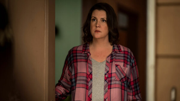 A dark haired middle aged woman in red plaid shirt and grey t-shirt answers the door