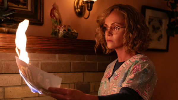 A woman with crimped blonde hair and glasses, dressed in a floral nurses top, burns a piece of paper