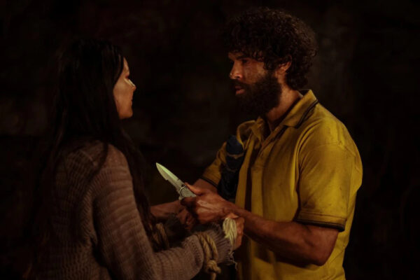 A dirty bearded dark haired man in a yellow collared shirt holds a knife near the chest of a brunette teen girl who clutches his wrists