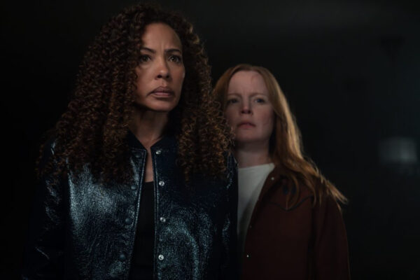 A redhead with a facial scar, wearing a rust coloured jacket over a white shirt, stands partially behind a Black woman wearing a black jacket