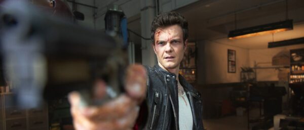 A brown haired man in a leather jacket points a gun directly at the camera