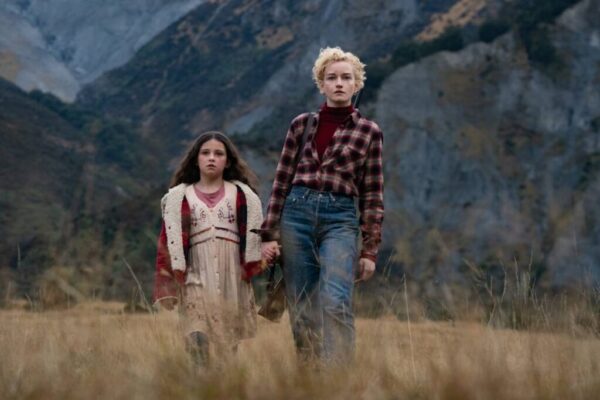 A blonde woman in a red flannel shirt (R) walks hand in hand with a young dark haired girl in a field with mountains in the background