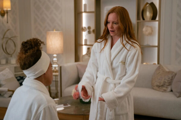 Two women - a red head (standing) and a Black woman (sitting) - talk while wearing white bath robes