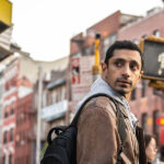 Riz Ahmed as Ash, wearing a grey hoodie, tan jacket and black backpack