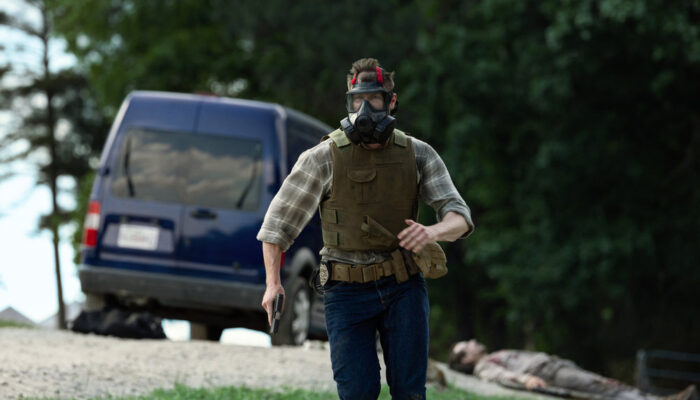 Olsen (Bill Heck) running away from a van, wearing body armor and a gas mask