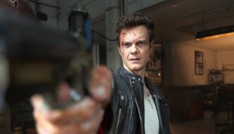 A brown haired man in a leather jacket points a gun directly at the camera